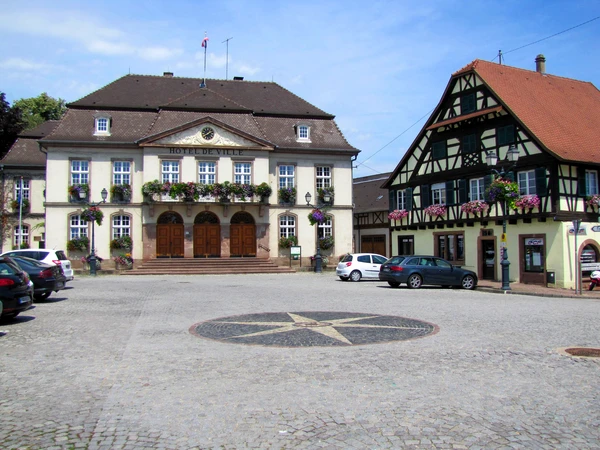Vue a&eacute;rienne ou paysage urbain d'Erstein montrant l'architecture typique alsacienne avec b&acirc;timents r&eacute;sidentiels et commerciaux, toitures traditionnelles, rue principale ou place de la ville avec quelques monuments ou b&acirc;timents caract&eacute;ristiques. L'image doit capturer l'essence d'une ville du Bas-Rhin, sans personnages, avec une qualit&eacute; photographique professionnelle.