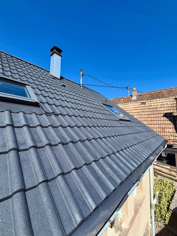 Photographie d'un ouvrier qualifié effectuant une inspection ou des travaux de réparation sur une toiture en tuiles ou ardoise, en toute sécurité avec harnais, situé sur les pentes du toit d'une maison résidentielle. La maison doit être clairement visible en arrière-plan, de style alsacien ou traditionnel. La lumière doit être naturelle et la scène doit transmettre professionnalisme, expertise technique et sérieux du travail de couverture.