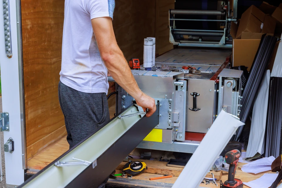 Un artisan qualifié opérant une machine à profiler mobile sur un chantier résidentiel, fabricant une gouttière aluminium en continu. La machine est installée directement sur la toiture ou à proximité immédiate de la maison. L'artisan porte des équipements de sécurité appropriés. On voit la bande d'aluminium en cours de profilage, la pièce en sortie de machine, et en arrière-plan une maison avec sa toiture. L'image capture le processus de fabrication précise et l'installation directe sur site qui caractérisent le service de l'entreprise.